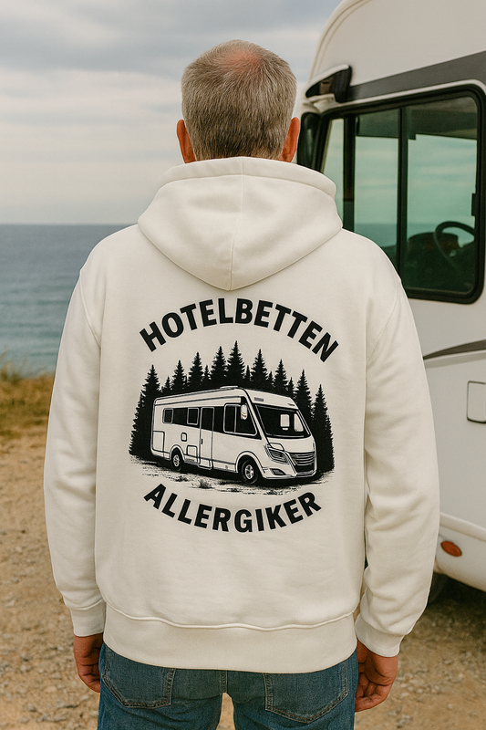 Ein Mann in weißem Hoodie von Camper4Love mit dem Aufdruck „Hotelbetten Allergiker“ und Wohnmobil-Motiv steht vor einem vollintegrierten Wohnmobil an der Küste – perfekt für Vanlife-Fans, Wohnmobilreisen und Outdoor-Abenteuer.