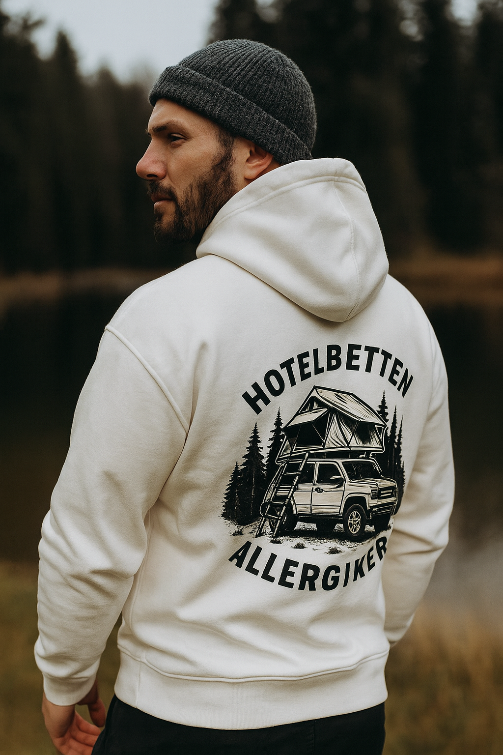 Mann trägt weißen Zipper Hoodie mit Aufdruck „Hotelbetten Allergiker“ und Camping-Motiv vor Waldkulisse – ideal für Outdoor- und Vanlife-Fans von Camper4Love.