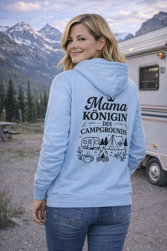 Frau trägt hellblauen Hoodie mit dem Design „Mama Königin des Campgrounds“ und schwarzem Camping-Motiv aus Wohnwagen, Zelt, Lagerfeuer und Natur, steht vor einem Wohnwagen in den Bergen