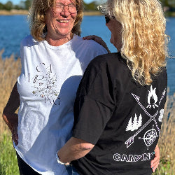 Zwei Frauen stehen am Wasser und lachen. Sie tragen T-shirts. Die eine Frau trägt ein weißes T-Shirt mit einem Vintage Aufdruck und die andere Frau trägt ein schwarzes T-Shirt mit dem Aufdruck Pfeilkreuz Wohnmobil